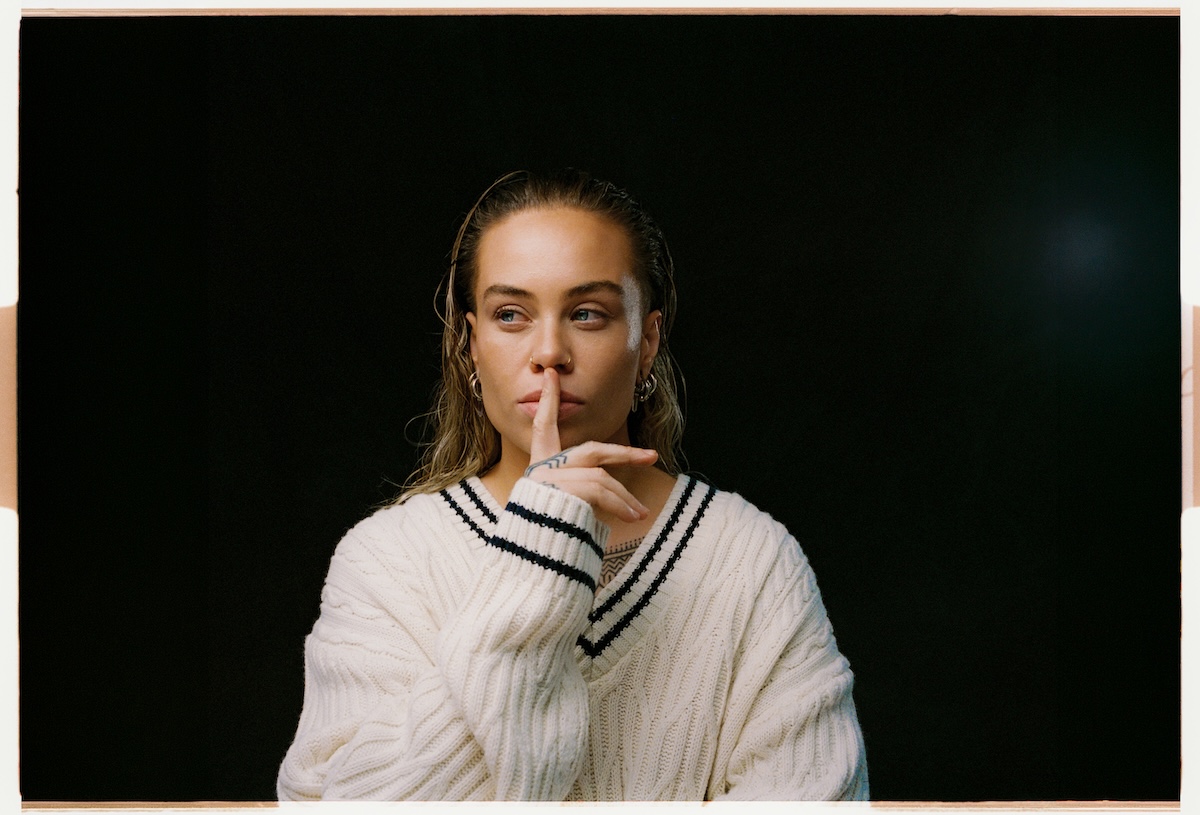 Australian musician Tash Sultana wearing a white sweater, holding a finger to their lips in a studio portrait photographed by Ben McFadyen.